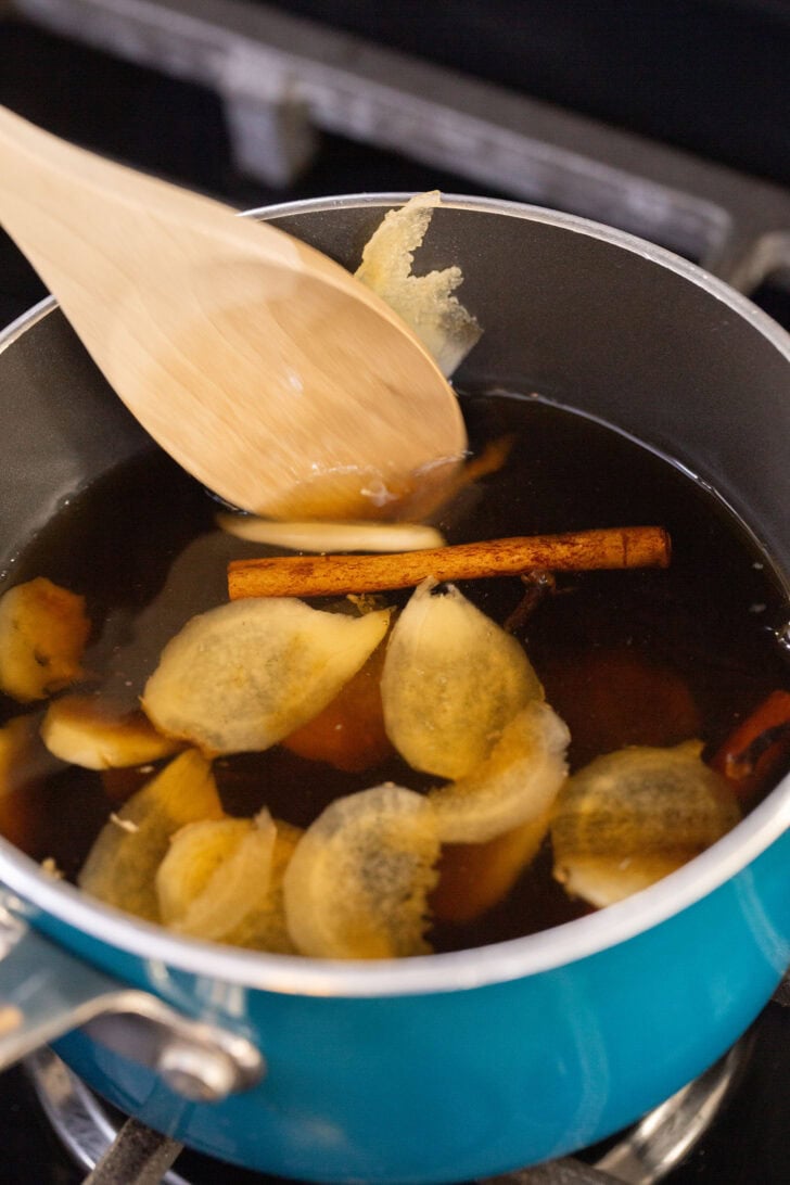 Stirring sliced ginger and a cinnamon stick into gingerbread simple syrup with a wooden spoon as it simmers on the stove.