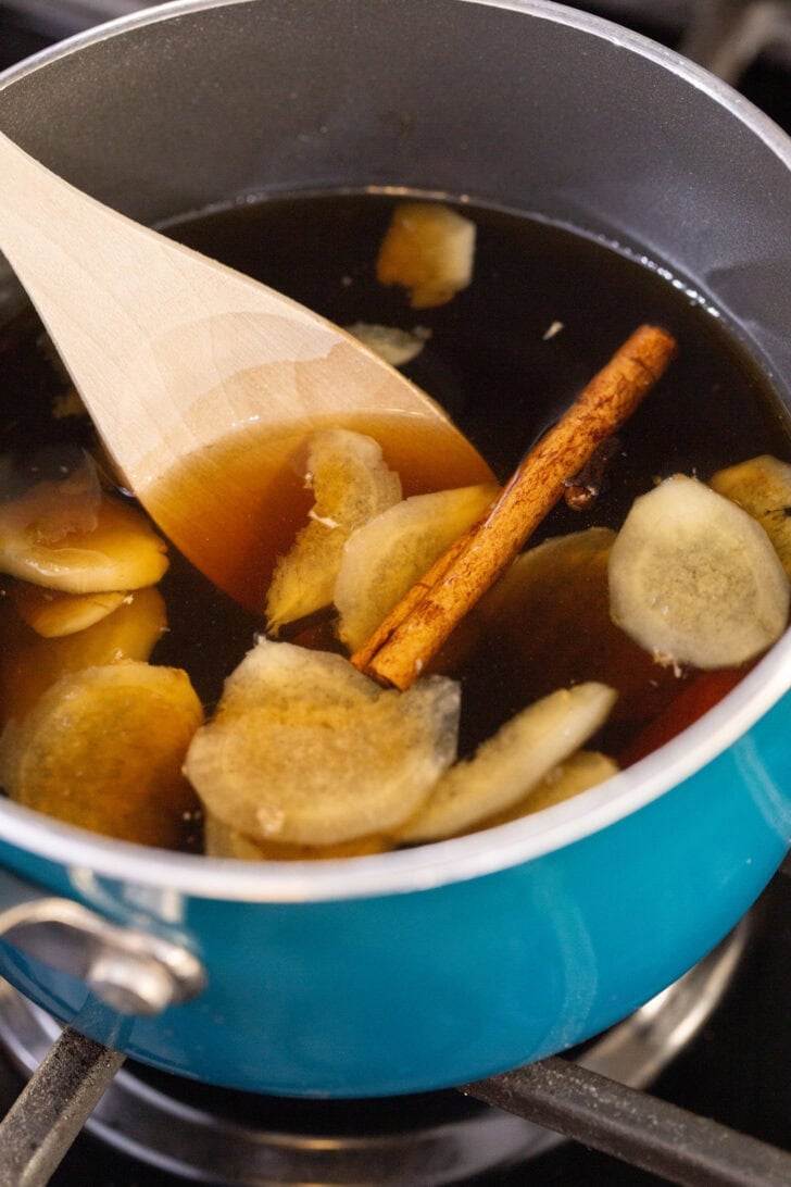 An overhead shot of gingerbread simple syrup steeping in a teal saucepan with ginger slices, whole cloves, and a cinnamon stick.