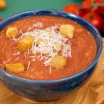 A side shot of 3-ingredient white bean tomato soup in a blue bowl on a cutting board, topped with croutons and parmesan cheese.