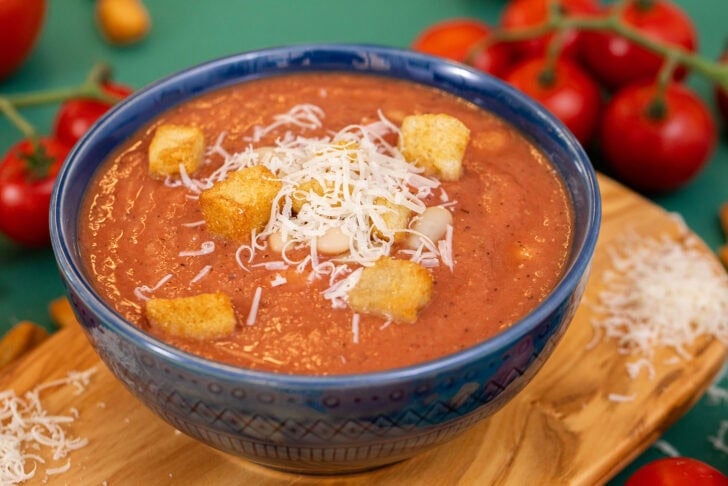 A side shot of 3-ingredient white bean tomato soup in a blue bowl on a cutting board, topped with croutons and parmesan cheese.