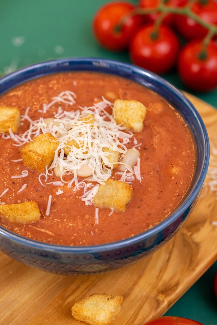 A close side shot of 3-ingredient white bean tomato soup in a blue bowl on a cutting board, topped with croutons and parmesan cheese.