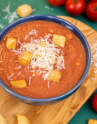 A close up shot of 3-ingredient white bean tomato soup in a blue bowl on a cutting board, topped with croutons and parmesan cheese.