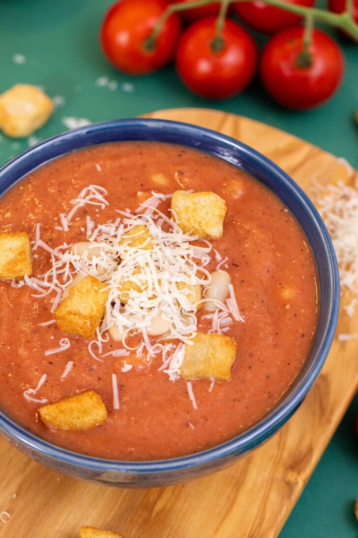 A very close up shot of 3-ingredient white bean tomato soup in a blue bowl on a cutting board, topped with croutons and parmesan cheese.