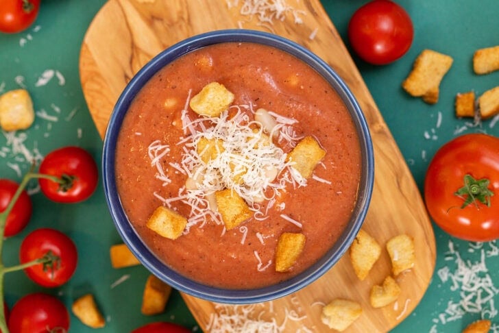An overhead shot of 3-ingredient white bean tomato soup in a blue bowl on a cutting board, topped with croutons and parmesan cheese.